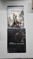 Two hardcover video game strategy guides on a white surface: Assassin's Creed III Prestige Edition and Call of Duty Modern Warfare 2 Official Strategy Guide.