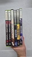 Front view of the Xbox 360 game cases held in hand showcasing titles Gears of War 2, Gears of War, Star Wars: The Force Unleashed, and multiple Mass Effect titles.