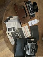 Overview photo showing Polaroid Land Camera in case, instruction manuals and Stereo Realist camera on table.