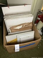 Box filled with various blank canvases of multiple sizes showing wooden frames and white painting surfaces. Some canvases wrapped in torn brown paper.