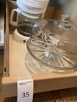Overview photo showing clear glass cake stand with dome next to white drip coffee maker in drawer