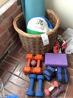 Photo showing two pairs of dumbbells (orange and blue), blue adjustable ankle weights, purple yoga block, blue-green rolled yoga mat inside a wicker basket, and a multicolor beach ball on top of the mat
