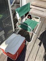 Photo of green folding chair with metal frame and green canvas seat and backrest alongside red and white cooler placed on a wooden deck near a glass door.