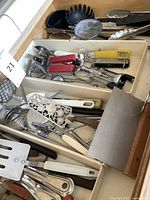 Photo showing a drawer with a variety of metal and plastic kitchen utensils including potato masher, tongs, bottle openers, strainer, and nutcracker.
