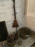 Photo showing large metal bucket filled with pinecones, metal pitcher, tea kettle, broom hanging on wall against white brick.