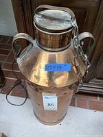 Full view of metal milk can showing its copper color, two side handles, attached lid and measurement tag.