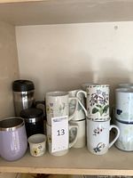 Shelf with various mugs, including stainless steel travel mugs and ceramic mugs with floral patterns, along with a small ceramic cup and a light purple metal travel mug.