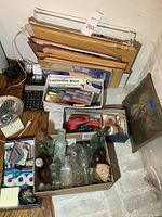 Photo showing office supplies including calculator, mailing envelopes, pens, stapler, small fan, tape rolls, and a box with antique glass bottles next to a framed metal art piece.