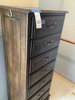 Side view of the tall dresser showing six drawers with ornate metal handles and worn wooden finish.