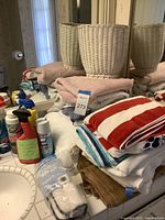 Photo showing stacked assorted towels, white wicker waste basket behind them, and several cleaning supply bottles around them on a bathroom counter.