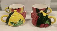 Two hand-painted teapots with matching cups, showing both items with colorful fruit and leaf designs.