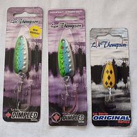 Three Len Thompson fishing lures in original packaging, two from the Dimpled series and one from the Original series, displayed on light fabric background.