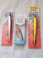 Photo showing three Rebel fishing lures: two in original packaging (white/silver and yellow/gold) and one POP-R lure in box