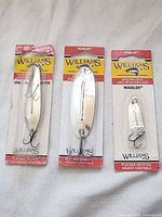 Photo showing three Williams fishing lures in their original packaging, including two ice jig spoon lures and one smaller hammered Wabler spoon lure.