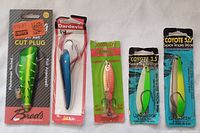 Photo showing four distinct fishing lures in their original packaging: one green cut plug, one blue feathered spoon, one copper colored casting lure, and two salmon trolling spoons.