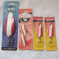Four packaged fishing lures on light background; one Blue Fox brand blue card backing, one ETIC branded pink card, and two Red Wolf branded yellow cards. All lures have red and white coloring with treble hooks.