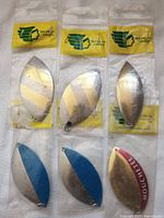 Six metallic fishing lure blades, three silver and gold striped in Majik Canada bags, two blue/gold, one gold with red branding.