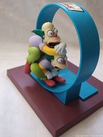 Side view of Krusty and Clown Homer figures stacked on a blue circular base with a brown rectangular platform beneath.