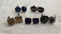Seven pairs of cufflinks shown on white fabric background, various designs and colors.