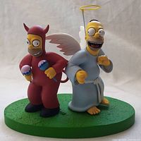 Front view showing both Homer Simpson figurines: devil Homer holding maracas and angel Homer with halo, wings, and robe standing on green textured base.