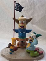 Three Simpsons figures displayed: Bart Simpson, Krusty the Clown kneeling, and a totem pole with 'Camp Bart' flag on a sandy base.