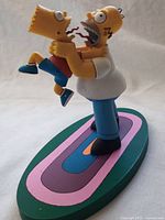Side angle showing Homer Simpson grasping Bart Simpson by the neck with both characters on an oval colorful base.