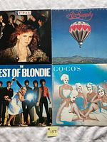 Top view of four vinyl album covers: T'Pau, Air Supply, Blondie, The Go-Go's arranged on white cloth.