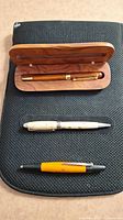 Three pens displayed: top is medium-toned wood pen in wooden case, middle is cream wood pen with silver accent, bottom is orange-toned wood Faber-Castell pen.