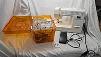Brother XL-3022 white sewing machine with black foot pedal and amber storage case next to it with compartments for notions.