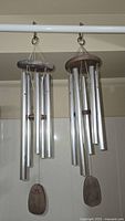 Front view of two metallic tube chimes with wooden tops and windcatchers hanging on a white bar.