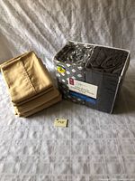 Two double bed sheet sets on a bed, one in a clear plastic package with geometric pattern and solid gray sheets visible, labeled Mainstays 2-pack cotton sheets, and one tan-colored folded sheet set beside it.