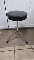 Front view of black round padded drum throne with adjustable height and tripod legs.