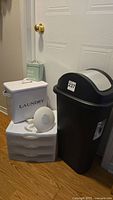 Photo of the lot showing all included items: black trash receptacle, white enamel laundry soap box with damage, green tin lint bin, white storage drawers, and round alarm clock.