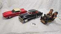 Two tin friction cars, a black 1950 Cadillac Sedan and a red 1963 Ford Thunderbird, with a small figurine of race car drivers, all displayed on white cloth.