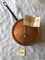 Copper saucepan with lid placed on a white tablecloth. Pan diameter about 6.5 inches with a long handle. LifeStyle paper tag attached to the lid handle.
