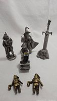 Five mystical pewter figures arranged against white cloth background: knight, wizard Merlin, sword in stone, dwarf couple, and a figure 'The Evil of Greed'.
