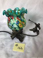 Lamp with multicoloured stained glass shade shaped like a ruffled waterlily, seen alongside attached dark metal cupid figurine playing trumpet on leaf platform and metal leaf-shaped base.
