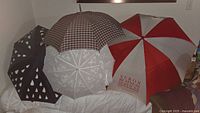 Four umbrellas displayed together: a black umbrella with raindrop design, a white lace wedding parasol with scalloped edges, a red and white promotional umbrella, and a plaid umbrella, showing their tops and details.