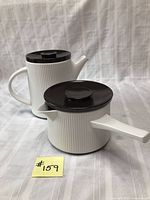 Full view of both cookware pieces showing the white ribbed ceramic and contrasting dark brown lids, set on a white textured cloth.