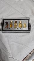 Front view of the sealed box containing five miniature perfume bottles arranged in a line within a clear plastic case with black and white background labeled '5 Parfums de Paris'.