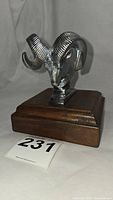 Front angled view of the chrome ram hood ornament on a wood base showing texture and shine of metal and wood finish.