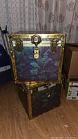 Two stacked metal trunks with purple and teal fern leaf pattern and brass accents, showing general aged condition.