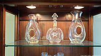 Shelf display of all three wine decanters: two have handle grips with lip pour, one is Louis XIII Remy Martin decanter with stopper.