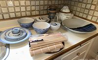 Overview of Asian themed ceramic serve ware on kitchen counter showing bowls, plates, jars, chopsticks, and mats.