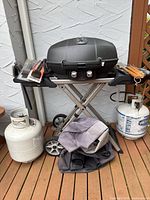 Full view of grill on folding stand with side shelves, two propane tanks on deck, utensils on side shelf and folded cover beneath