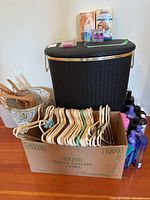 Overall view of items including laundry hamper, wicker basket, cardboard box of hangers, assorted hygiene product bottles, and boxed hair kits on top of hamper.