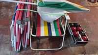 Photo showing five colorful striped fabric folding chairs stacked together with a folded green, yellow, white umbrella on top, all on a concrete floor.