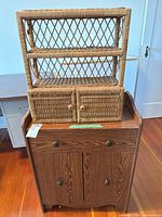 Photo of a wooden cabinet with drawer and double doors topped with a wicker hanging shelf with two open shelves and a small cabinet with woven doors.