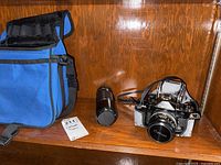 Blue camera carrying bag, Canon A-1 camera with lens attached, and telephoto lens placed on wood shelf
