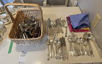 Overview of woven basket filled with mixed cutlery including spoons, knives and forks, plus laid out cutlery and folded linens on table.